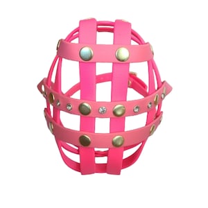 May include: A bright neon pink basket-style muzzle constructed from interconnected straps. The design features metallic gold-tone circular studs and small clear crystals embedded along the horizontal bands, creating a decorative grid pattern.