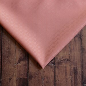 May include: A close-up of a salmon-colored fabric with a subtle textured pattern. The fabric is folded in half and placed on a brown wooden surface.