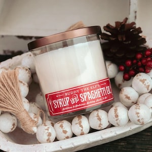 May include: A clear glass candle jar with a copper lid. The candle is white and has a red label that reads "Buddy the Elf's Syrup and Spaghetti Co. Nutritious & Delicious Holiday Meals To Go!"
