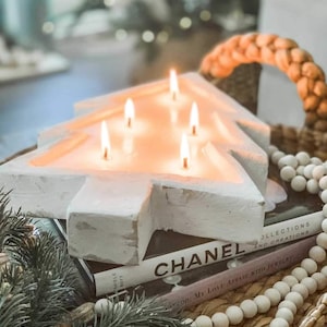 May include: A white ceramic Christmas tree candle holder with five lit candles. The candle holder is sitting on a stack of books, including one with the word "CHANEL" on the cover. A strand of white wooden beads is in the foreground.
