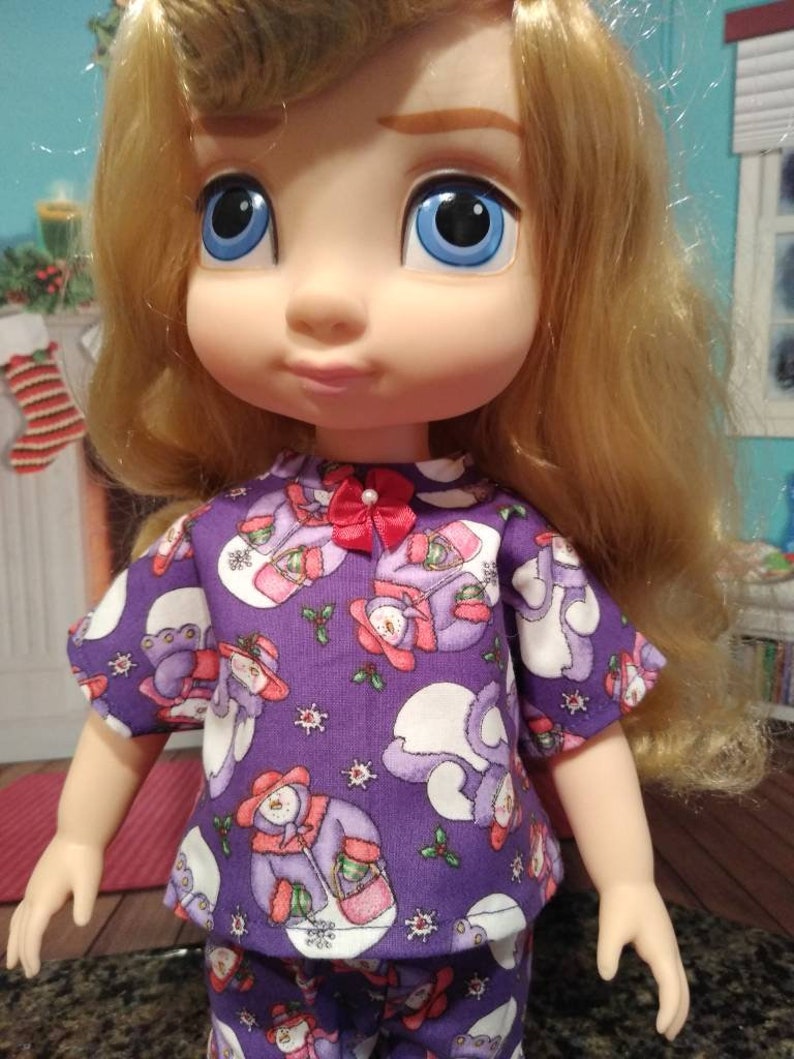 Może przedstawiać: Lalka w fioletowej i białej piżamie z wzorem bałwanka. Lalka ma długie blond włosy i niebieskie oczy. Piżama ma czerwoną kokardkę.