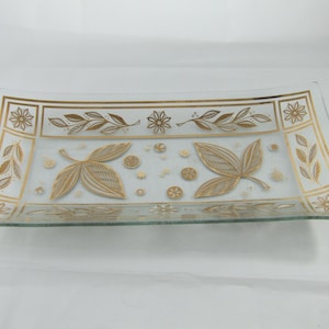 May include: Rectangular glass serving tray with gold leaf and floral designs. The tray features a clear glass base with gold accents, including stylized leaves, flowers, and geometric borders. The design is symmetrical, with the gold elements contrasting against the clear glass.