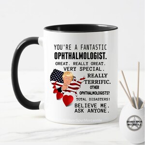 Pu&ograve; includere: Una tazza da caff&egrave; bianca con un bordo e un manico neri. La tazza presenta un'immagine di cartone animato di Donald Trump che punta verso lo spettatore. Il testo sulla tazza recita "You're a fantastic Ophthalmologist. Great. Really Great. Very Special. Really Terrific. Other Ophthalmologists? Total Disasters! Believe Me. Ask Anyone."