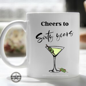 May include: White ceramic mug with a black design that says "Cheers to Sixty Years" with a martini glass and olives. The mug has a black handle.