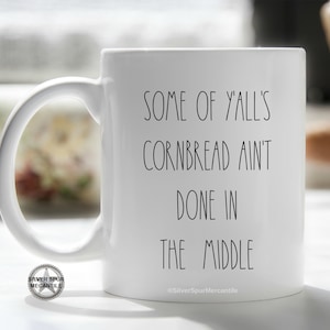 May include: White ceramic mug with black text that reads "Some of y'all's cornbread ain't done in the middle". The mug has a black and white logo on the bottom that reads "Silver Spur Mercantile".