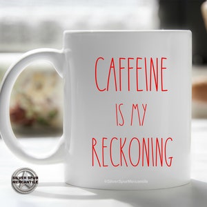 May include: White ceramic coffee mug with the red text "CAFFEINE IS MY RECKONING". The mug has a large handle and a smooth surface. The words are in a simple, sans-serif font. The mug is a standard size.