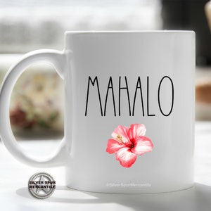 Puede incluir: Taza de caf&eacute; de cer&aacute;mica blanca con un contorno negro de la palabra "Mahalo" y un gr&aacute;fico de flor de hibisco rosa. La taza tiene un asa plateada.