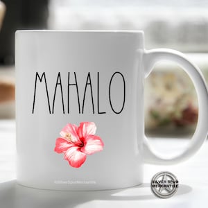 Puede incluir: Taza de cer&aacute;mica blanca con la palabra "MAHALO" en letras negras y un gr&aacute;fico de flor de hibisco rosa. La taza tiene un asa negra.