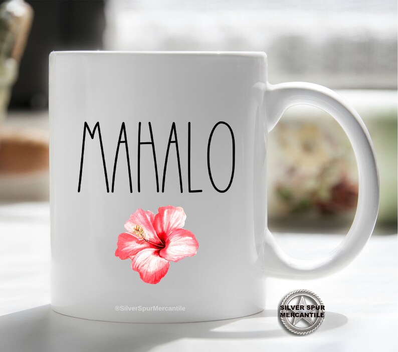 Puede incluir: Taza de cer&aacute;mica blanca con la palabra "MAHALO" en letras negras y un gr&aacute;fico de flor de hibisco rosa. La taza tiene un asa negra.