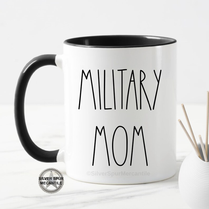 May include: White ceramic coffee mug with a black handle and black rim. The mug has the text "MILITARY MOM" printed in black on the front. The mug is sitting on a white surface.