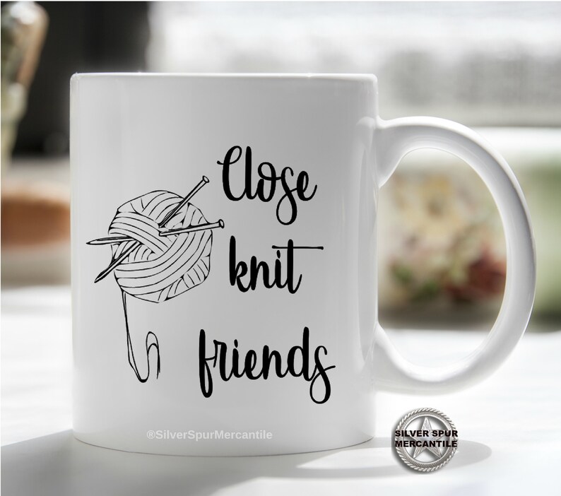 Może przedstawiać: Biały kubek ceramiczny z czarno-białym rysunkiem wł&oacute;czki i drut&oacute;w do rob&oacute;tek na drutach. Na kubku nadrukowany jest tekst "Close knit friends".
