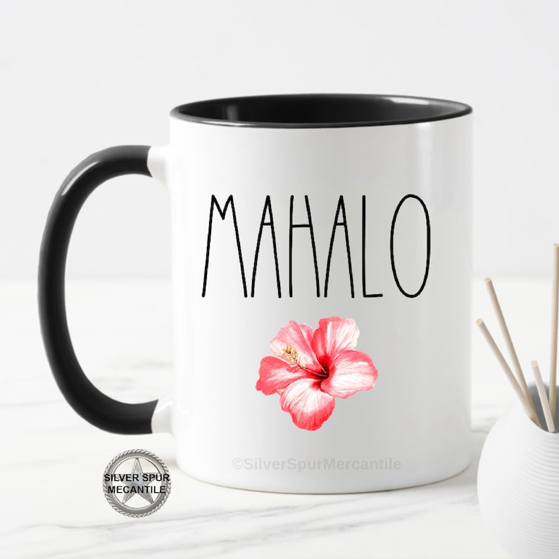 Puede incluir: Taza de caf&eacute; de cer&aacute;mica blanca con asa y borde negros. La taza tiene una inscripci&oacute;n negra que dice "MAHALO" y un gr&aacute;fico de flor de hibisco rosa. La taza est&aacute; sobre una superficie blanca.