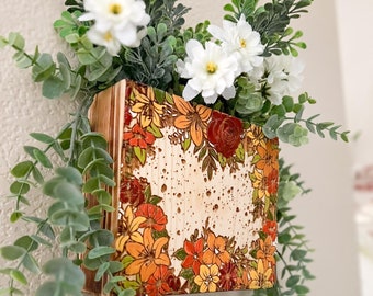 Op de afbeelding: Een kleine houten plantenbak met een bloemmotief dat op de voorkant is geschilderd. De plantenbak is gevuld met groen gebladerte en witte bloemen.