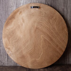 May include: Back of a round, light brown wooden board with a natural wood grain pattern. A metal sawtooth hanger is attached at the top center for wall mounting. The board is likely for a decorative item.
