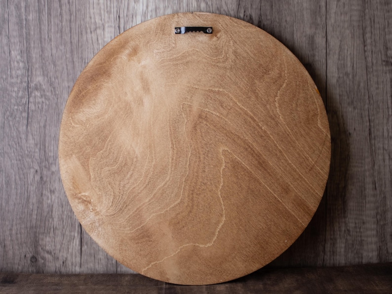 May include: Back of a round, light brown wooden board with a natural wood grain pattern. A metal sawtooth hanger is attached at the top center for wall mounting. The board is likely for a decorative item.