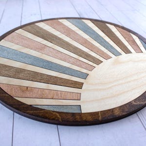 May include: A round wooden decorative piece with a sunburst design. The sunburst features alternating stripes of light and dark wood in shades of brown, pink, and blue. The piece is approximately 12 inches in diameter with a dark brown border.