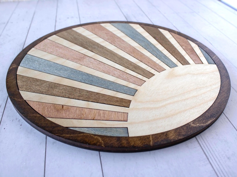 May include: A round wooden decorative piece with a sunburst design. The sunburst features alternating stripes of light and dark wood in shades of brown, pink, and blue. The piece is approximately 12 inches in diameter with a dark brown border.