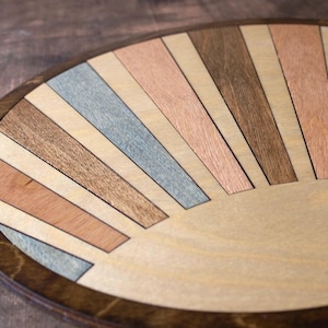 May include: A round wooden tray with a sunburst design. The tray features a dark brown rim and a light wood center. The sunburst pattern is made of various wood tones, including light brown, gray, and reddish-brown.