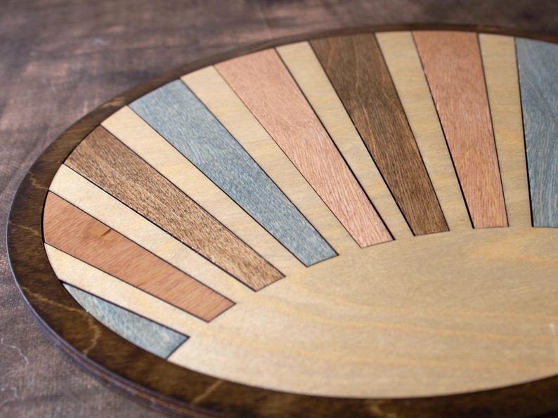 May include: A round wooden tray with a sunburst design. The tray features a dark brown rim and a light wood center. The sunburst pattern is made of various wood tones, including light brown, gray, and reddish-brown.