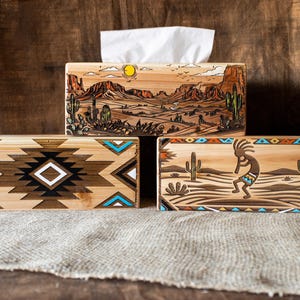 May include: Three rectangular wooden tissue box covers featuring engraved and painted designs. One displays a desert landscape with a sun, another a geometric diamond pattern in black, white, and turquoise, and the third a flute player with cacti.