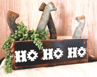 May include: A rustic wooden box with a dark stain, decorated with white geometric patterns. The box holds several weathered horseshoes and faux greenery. The background is a light wood panel.