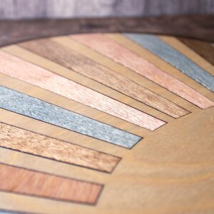 May include: Close-up of a wooden surface with a sunburst design. The design features alternating strips of wood in various colors, including light brown, copper, gray, and dark brown. The wood grain is visible, adding texture to the piece.