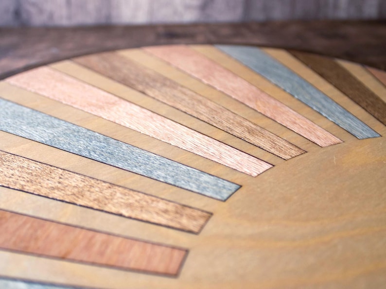 May include: Close-up of a wooden surface with a sunburst design. The design features alternating strips of wood in various colors, including light brown, copper, gray, and dark brown. The wood grain is visible, adding texture to the piece.