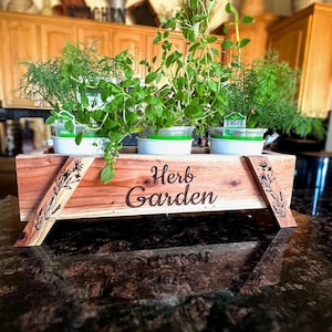 May include: A wooden planter box with the words "Herb Garden" engraved on the front. The planter is filled with four potted herbs, including dill, oregano, and thyme. The planter is sitting on a dark countertop.