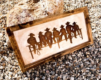 Op de afbeelding: Een ingelijst houten bord met een silhouet van zeven cowboys die op een hek zitten. Het bord heeft een rustiek bruin houten frame en een lichtbruine achtergrond.