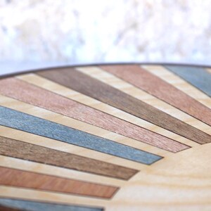 May include: Close-up of a round wooden surface with a sunburst design. The design features alternating stripes of light wood, copper, brown, and blue-gray tones, creating a geometric pattern. The edge of the surface is a dark brown.