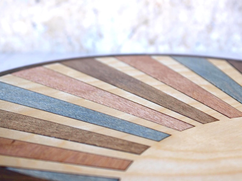 May include: Close-up of a round wooden surface with a sunburst design. The design features alternating stripes of light wood, copper, brown, and blue-gray tones, creating a geometric pattern. The edge of the surface is a dark brown.