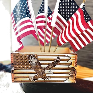 May include: Wooden box with an American flag design and an eagle carving. Small American flags on sticks are displayed inside the box. The flags have red and white stripes and blue fields with white stars.