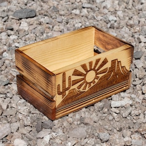 May include: A rectangular wooden crate with a desert landscape design. The design includes a sunburst, mountains, and a cactus, all etched into the wood. The crate has a natural wood color with darker, burnt edges.