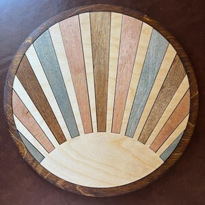May include: A round wooden wall art piece with a sunburst design. The sunburst is made of various wood tones, including light brown, gray, and pink. The piece has a dark brown, circular frame. The diameter is approximately 12 inches.
