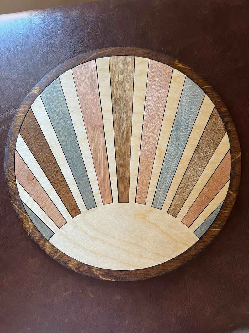 May include: A round wooden wall art piece with a sunburst design. The sunburst is made of various wood tones, including light brown, gray, and pink. The piece has a dark brown, circular frame. The diameter is approximately 12 inches.