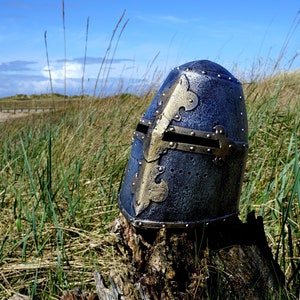 Op de afbeelding: Een zilveren en gouden ridderhelm met een kruisontwerp aan de voorkant. De helm staat op een boomstronk in een grasveld.