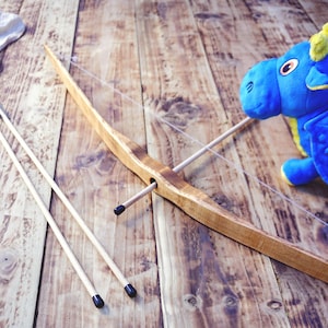 May include: A wooden bow with a clear string and two wooden arrows featuring black tips, accompanied by a blue plush dragon toy. The image includes text that reads Adventure Starts Here Handcrafted Made of wood in a dark green graphic.