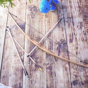 May include: A wooden toy bow and three arrows with black, blue, yellow, and green fletching. A blue plush dragon toy with yellow horns sits nearby. The text Adventure Starts Here Handcrafted Made of wood is visible in the upper left corner.