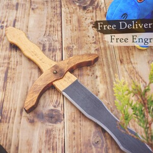 May include: A wooden toy sword with a light brown handle and a darker brown crossguard. The blade is a flat gray color. The sword is lying on a wooden surface.