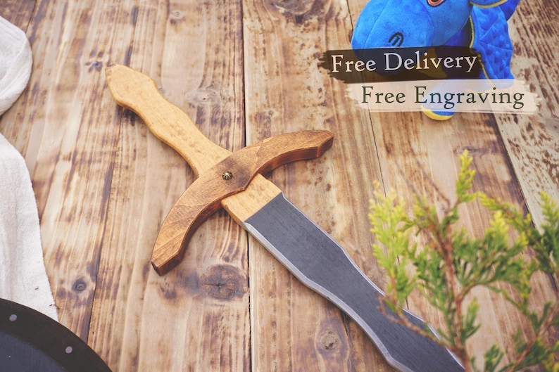 May include: A wooden toy sword with a light brown handle and a darker brown crossguard. The blade is a flat gray color. The sword is lying on a wooden surface.