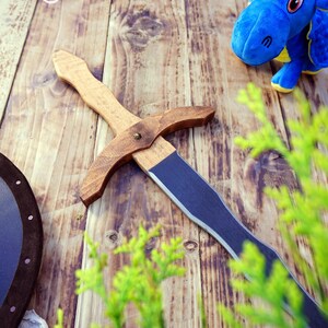 May include: A wooden toy sword with a dark blue blade and a light brown handle lies on a wooden surface. A blue stuffed dragon toy with yellow accents is in the background. A shield is also visible.