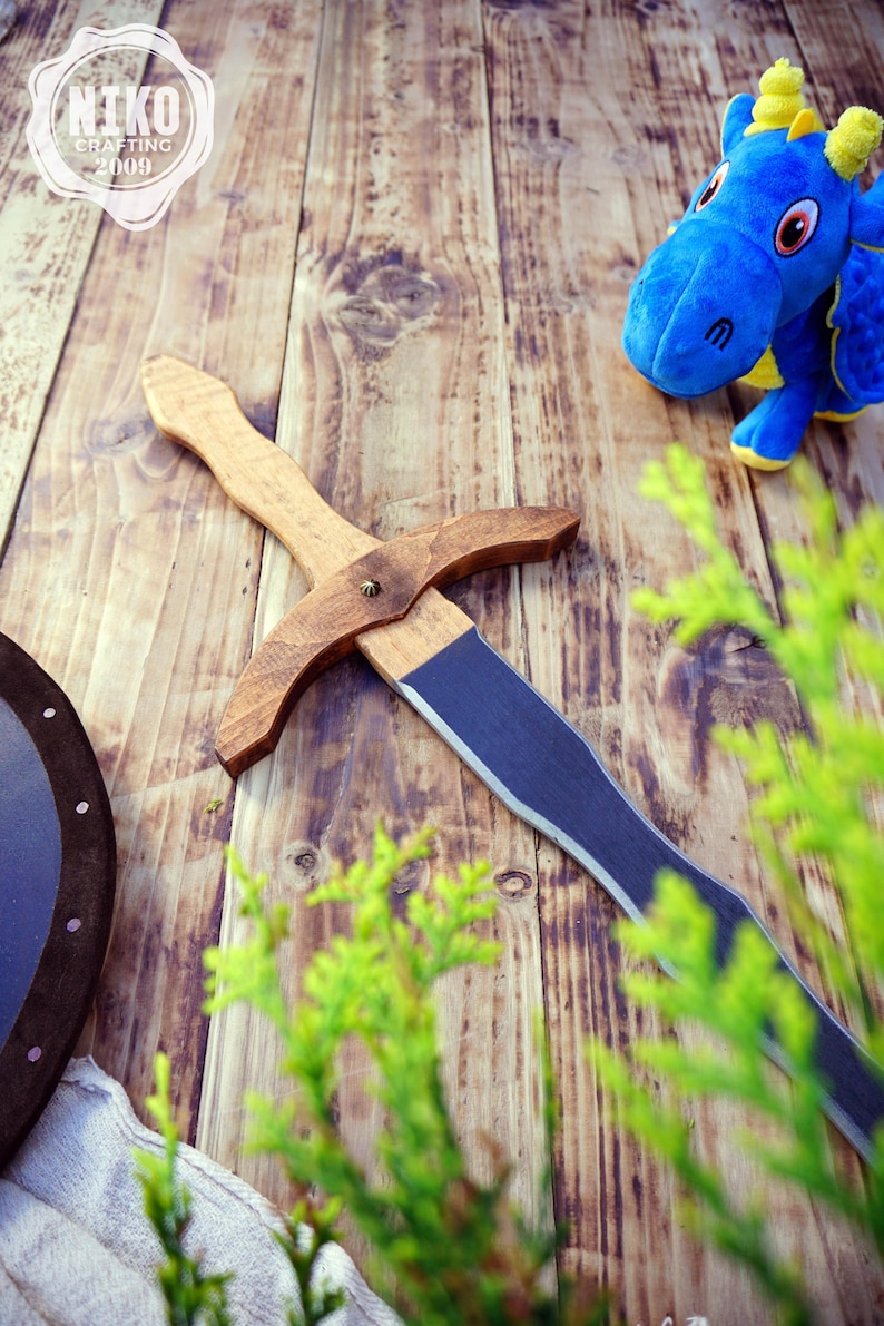 May include: A wooden toy sword with a dark blue blade and a light brown handle lies on a wooden surface. A blue stuffed dragon toy with yellow accents is in the background. A shield is also visible.