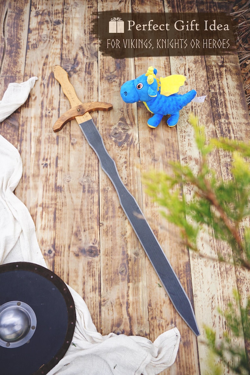 May include: A wooden sword with a gray blade and a brown handle lies on a wooden surface, next to a blue plush dragon with yellow wings. A black shield and the text "Perfect Gift Idea For Vikings, Knights Or Heroes" are also visible.