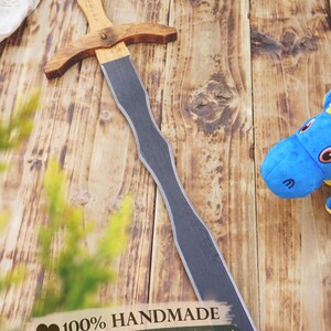 May include: A handmade wooden sword with a natural wood handle and a gray blade. The sword is lying on a wooden surface. The text "100% HANDMADE" is visible.