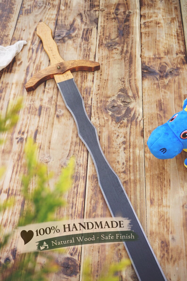 May include: A handmade wooden sword with a natural wood handle and a gray blade. The sword is lying on a wooden surface. The text "100% HANDMADE" is visible.