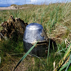 Op de afbeelding: Een zilveren en gouden ridderhelm met vizier, liggend in hoog groen gras.