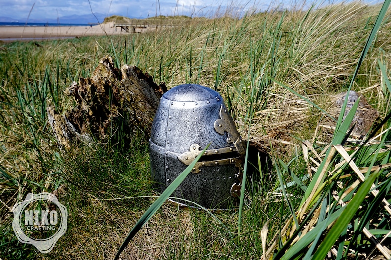 Op de afbeelding: Een zilveren en gouden ridderhelm met vizier, liggend in hoog groen gras.