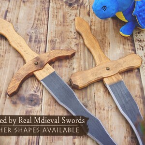 May include: Two wooden toy swords with light brown handles and gray blades rest on a wooden surface. The text "Inspired by Real Medieval Swords" and "OTHER SHAPES AVAILABLE" is visible. A blue stuffed dragon toy is in the background.