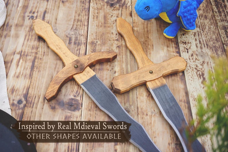 May include: Two wooden toy swords with light brown handles and gray blades rest on a wooden surface. The text "Inspired by Real Medieval Swords" and "OTHER SHAPES AVAILABLE" is visible. A blue stuffed dragon toy is in the background.