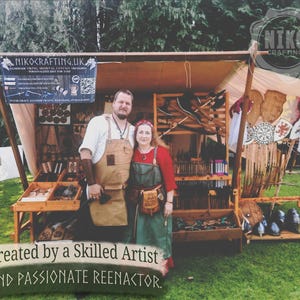 May include: A vendor booth displaying handmade Viking, medieval, and fantasy treasures. The booth features a variety of leather crafts, wooden items, and weaponry. The sign reads "NIKOCRAFTING.UK" and "Created by a Skilled Artist and Passionate Reenactor."